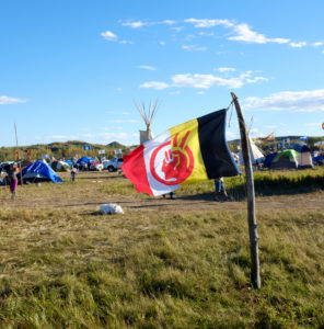 The American Indian Movement flag is a common sight at the camps. Thick vertical bars of red, white, yellow, and black background a red face that is also a raised fist and a peace sign. It can be seen atop flag poles, off the back of pickup trucks, or at individual camps as seen here. Image by author, September 2016.