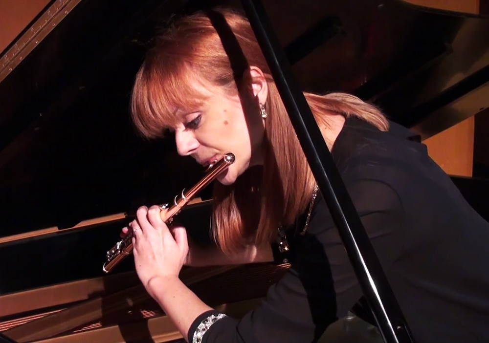 A woman plays the flute while crouched beneath the propped cover a grand piano.