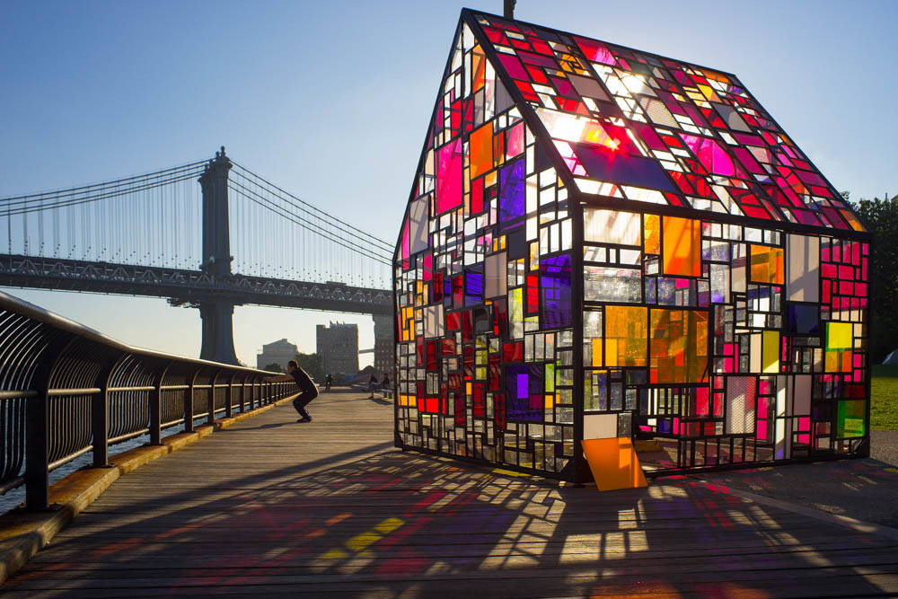 A small house with a multi-colored stained glass exterior. The Brooklyn Bridge in the background.