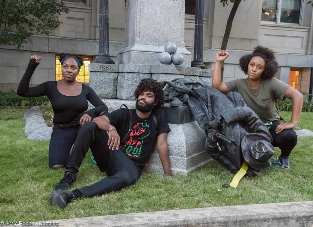 Three young people gather around a bronze statue, lying on its side, and raise their fists.