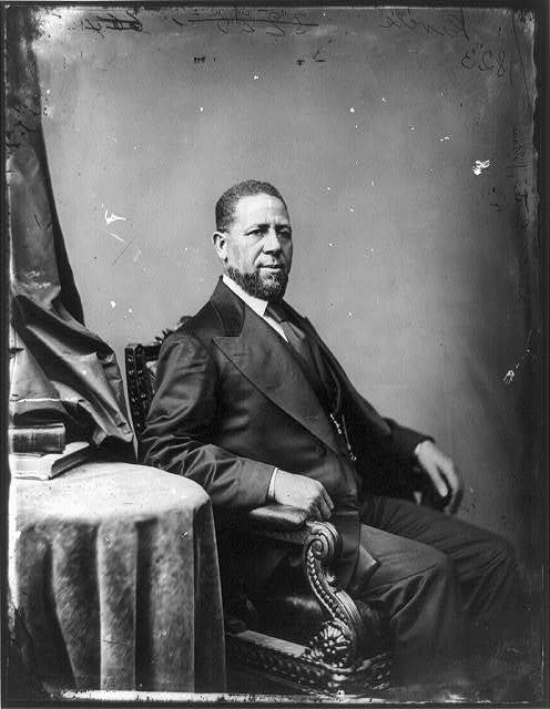 A man sits in formal dress next to a small table and a flag.