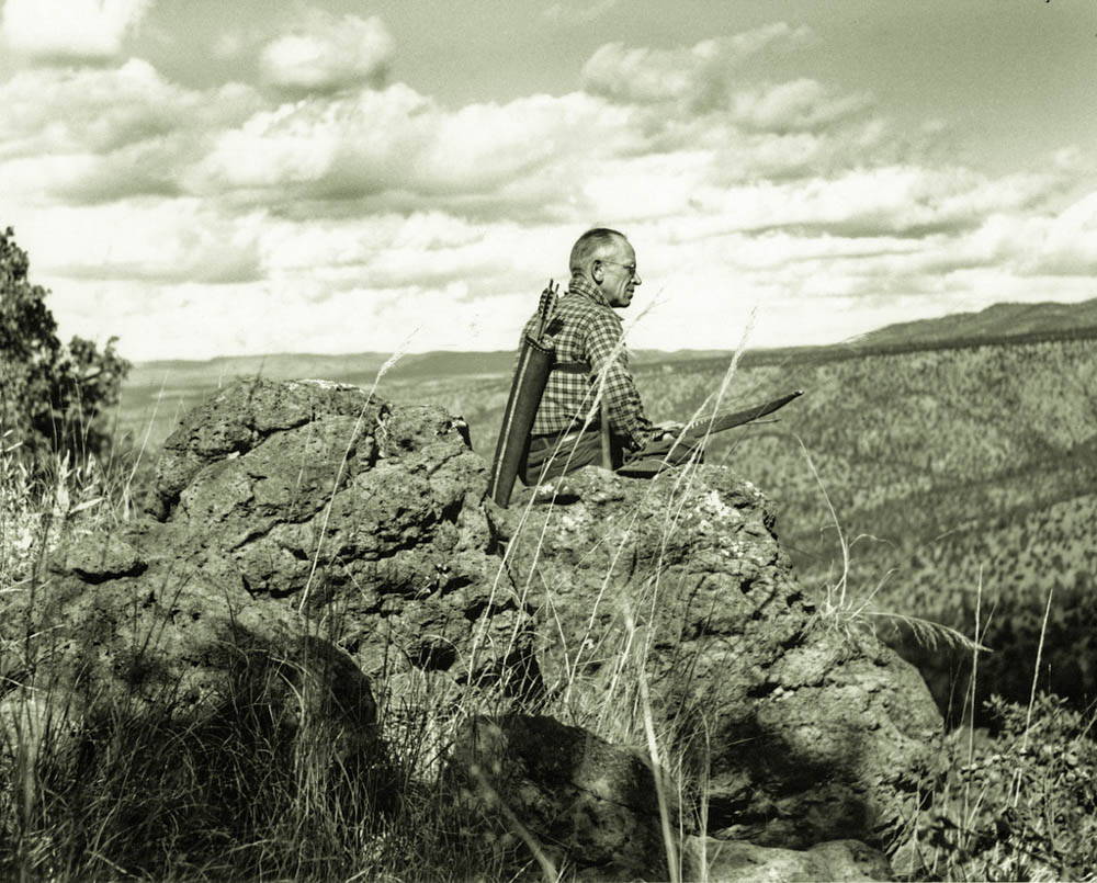 A man holding a bow with a quiver of arrows on his back sits on a rocky outcorop looking down over a expansive vista.