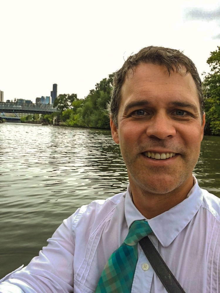 A man in a blue and green necktie poses for a selfie in the middle of a river with an urban skyline in the background.
