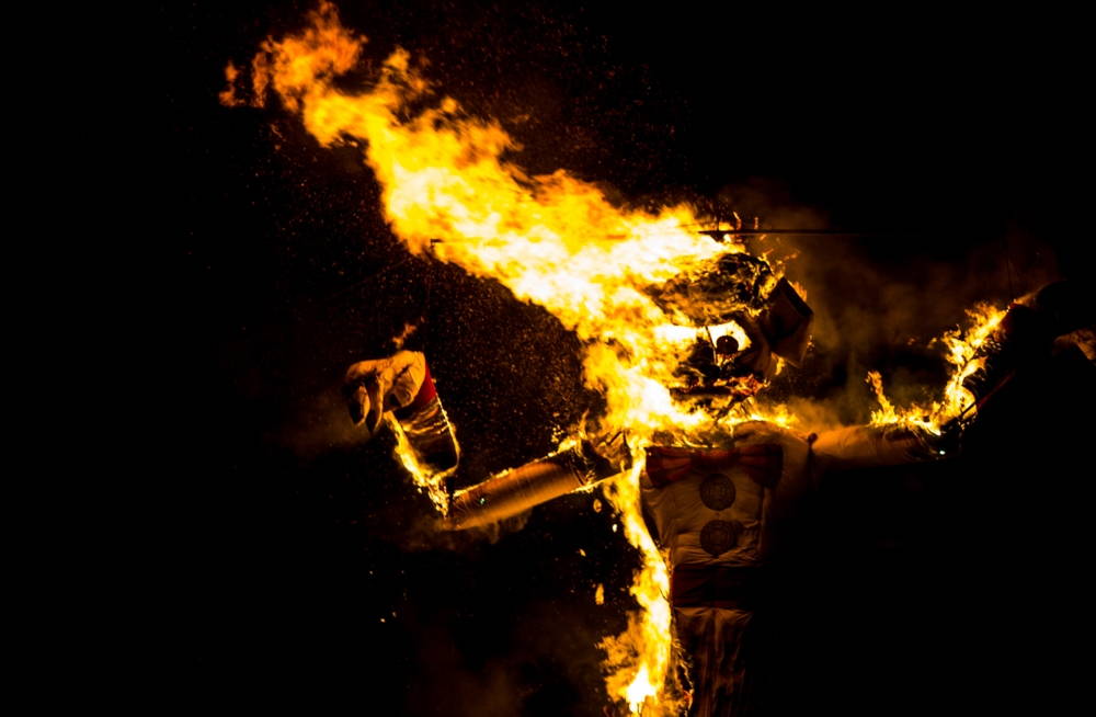 Zozobra, a 50-foot effigy, with its arms raised burns against a black background.