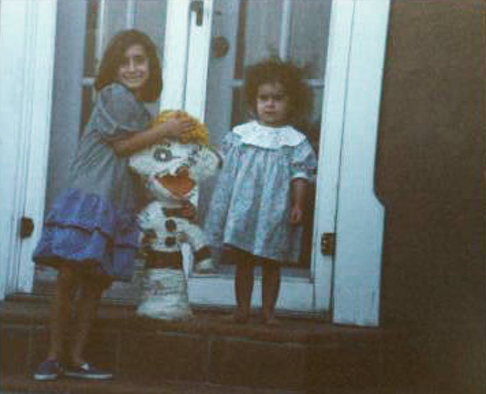 Two young girls in blue dresses stand outside a house holding up a large white Zozobra pinata.