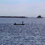 Ryan Griffis, Santarem, Brazil, 2016. Where the Tapajos and Amazon Rivers meet, a fleet of barges serves a nearby Cargill soy-loading facility. Brazil’s Cerrado biome and its lifeways have been decimated by increased soy production. Two fisherman in a small boat appear in the foreground. Large barges are visible floating across the horizon.