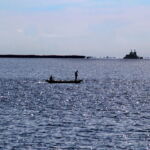 Ryan Griffis, Santarem, Brazil, 2016. Where the Tapajos and Amazon Rivers meet, a fleet of barges serves a nearby Cargill soy-loading facility. Brazil’s Cerrado biome and its lifeways have been decimated by increased soy production. Two fisherman in a small boat appear in the foreground. Large barges are visible floating across the horizon.