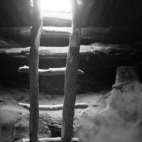 A ladder ascends from the inside of a kiva towards the daylight above. A black and white photograph of a ladder descending from a rooftop entrance.