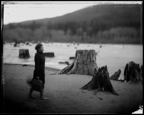Tomiko Jones. From Rattlesnake Lake, 2013. Platinotypes (from scanned 4x5 film negatives) 16x20 A woman wearing a coat and hat walks across a beach carrying a briefcase. Tree stumps are scattered across the beach and there is a mountain in the background.