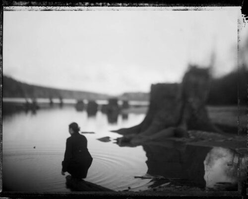Tomiko Jones. From Rattlesnake Lake, 2013. Platinotypes (from scanned 4x5 film negatives) 16x20 A woman carying a bag walks into the water. In the background are many treestumps and sky.