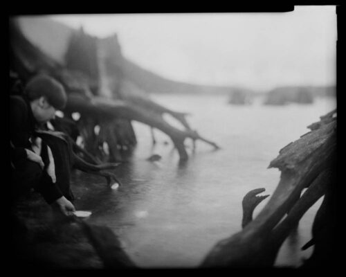 Tomiko Jones. From Rattlesnake Lake, 2013. Platinotypes (from scanned 4x5 film negatives) 16x20 A person on the bank of the water appears to be placing a piece of white paper onto the water. There are tree roots in the foreground and background.