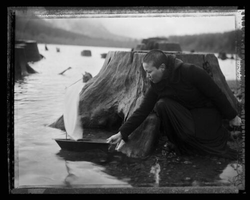 Tomiko Jones. From Rattlesnake Lake, 2013. Platinotypes (from scanned 4x5 film negatives) 16x20 A person kneels next to a tree stump at the water's edge. They push a small wooden boat with a white sail into the water.