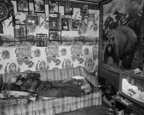 Tom Jones. Choka Watching Oprah, 1998. Silver gelatin print. An older man lays across a stripped couch. He pulls up his glasses to see Oprah on the television across the room. A wall hanging of a brown bear looms larger than the television.