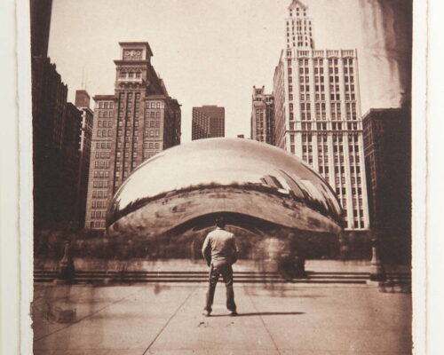 Eric Baillies. "The Bean." Salt print. 8x10 The sklyline of Chicago fills the background. In the mid-ground the Bean sculputures is centered. A man stands facing away from the camera and toward the Bean in the foreground.