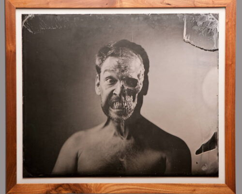 Eric Baillies. "The Machine." Double-exposed tintype. 20x24 Black and white photograph of a man from his torso up. Half of his face appears to be decomposed to the bone.