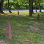 Public Soil Memory for the Plantationocene. Image by Raina Martens, 2017. Three pyramid-shaped stones rise out from green grass in a wooded lot. Public Soil Memory exhibit.