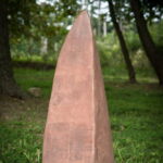 Stela imprinted with Richard Thomas Sr.’s probate records, listing the names of people who were enslaved by him. Image by Pete Duvall, 2017. A rust-colored stone rises from a green field. Public Soil Memory exhibit.
