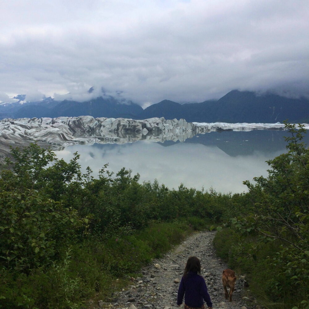 Girl and Dog on Glacier's Fringe, Eyak, Alaska. Photo by author. Gravel walkway and grass in foreground. Glacier and mountain range in the background.