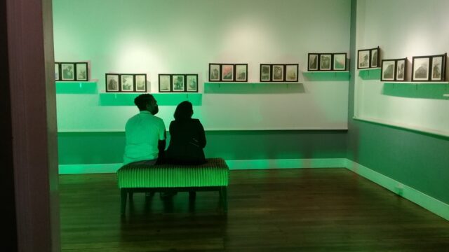 Installation of triptychs in Quiet as drawings exhibit. Photograph by David Cave, 2021. Two people sitting on a bench and looking at drawings in photo frames placed on shelves