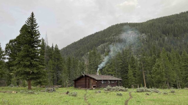 Cabin where forest service workers stay while on the job. Photo by the author, 2021 Brown cabin with smoke coming from the chimney and pine tree at the base of a mountain