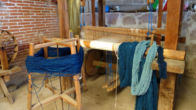 Loom at Santiago Ixtaltepec. Santiago Ixtaltepec is a village located about 15 kilometers east of Oaxaca City, just off the main highway. With the decline in agriculture in this region, many of the residents now specialize in one or another stage of wool production, from pasturing sheep to spinning and dying wool to weaving rugs and wall hangings for sale to shops in Oaxaca City. Such operations bring in much-needed cash to purchase household goods, tying these areas increasingly into urban and even global economies. The loom in the image above is one of three set up in the family's courtyard. A large loom with several large bundles of yarn in several shades of blue.