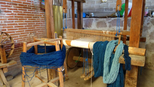 Loom at Santiago Ixtaltepec. Santiago Ixtaltepec is a village located about 15 kilometers east of Oaxaca City, just off the main highway. With the decline in agriculture in this region, many of the residents now specialize in one or another stage of wool production, from pasturing sheep to spinning and dying wool to weaving rugs and wall hangings for sale to shops in Oaxaca City. Such operations bring in much-needed cash to purchase household goods, tying these areas increasingly into urban and even global economies. The loom in the image above is one of three set up in the family's courtyard. A large loom with several large bundles of yarn in several shades of blue.