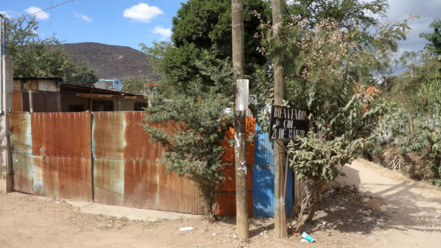 Colonia 21 de Marzo, Santa Cruz Xoxocotlán. On the one hand, the colonia is organized on a small grid with street signs, electric and telephone service, and other urban elements. On the other hand, roads remain largely unpaved, corrugated metal fencing and roofing is common, and houses are interspersed with open land. Many families in this colonia split their economic activity between local work as farm laborers and household producers (raising chickens especially) and jobs in Oaxaca City as drivers, kitchen workers, and street vendors. A corrugated metal fence forms the boundary of a house's compound along a dirt road