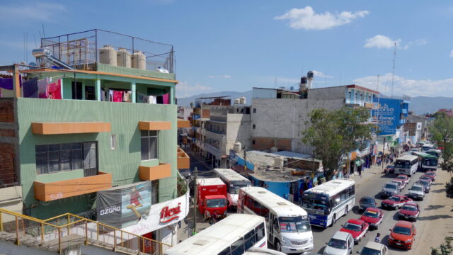 Heading into and out of Oaxaca. This image shows the crowded bus stage along the Periférico near the Central de Abastos (main wholesale market). Thousands of people converge here every day, traveling from villages, farms, and towns in order to shop for provisions in the city. Transportation stages and large markets play a crucial role in knitting together an urban system that extends well beyond the geographical boundaries of Oaxaca City. An intersection crowded with buses next to a green building.