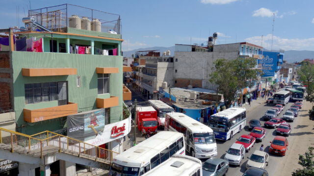 Heading into and out of Oaxaca. This image shows the crowded bus stage along the Periférico near the Central de Abastos (main wholesale market). Thousands of people converge here every day, traveling from villages, farms, and towns in order to shop for provisions in the city. Transportation stages and large markets play a crucial role in knitting together an urban system that extends well beyond the geographical boundaries of Oaxaca City. An intersection crowded with buses next to a green building.