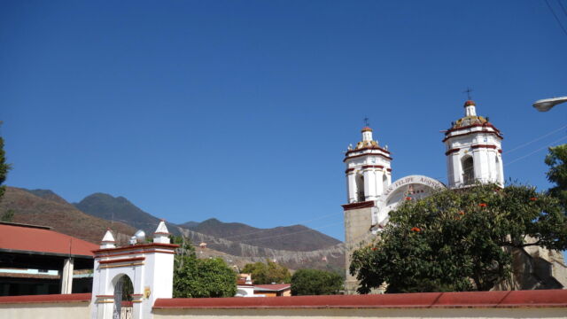 The Templo de San Felipe Apóstol. The old village of San Felipe del Agua presents a small remnant Spanish grid of a few blocks around the church plaza, while the rest of the village stretches out along either side of the road into Oaxaca City. The land surrounding the village was once primarily agricultural and pastoral, with a large ejido (communal land) devoted to the cultivation and harvesting of agave for mezcal production. Today, the land serves a mix of functions as the area has been incorporated into greater Oaxaca. A white and red church with a large fenced churchyard.
