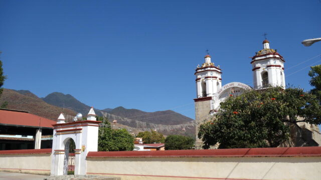 The Templo de San Felipe Apóstol. The old village of San Felipe del Agua presents a small remnant Spanish grid of a few blocks around the church plaza, while the rest of the village stretches out along either side of the road into Oaxaca City. The land surrounding the village was once primarily agricultural and pastoral, with a large ejido (communal land) devoted to the cultivation and harvesting of agave for mezcal production. Today, the land serves a mix of functions as the area has been incorporated into greater Oaxaca. A white and red church with a large fenced churchyard.