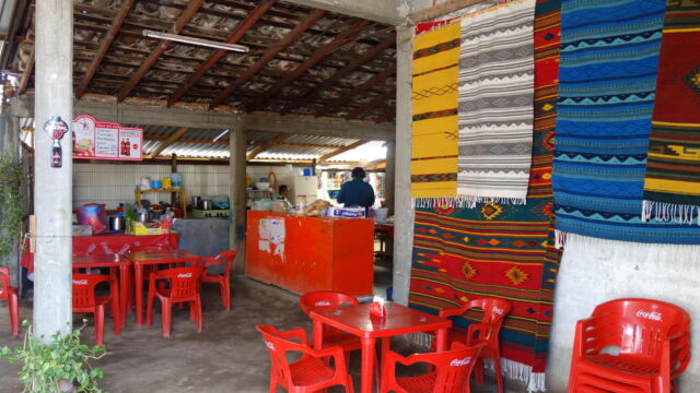 Restaurant, Teotitlán del Valle. This canteen-style restaurant is typical of peri-urban areas such as Teotitlán del Valle, a village located some 20 kilometers from the center of Oaxaca City. The restaurant is simply but elegantly designed, with an open-air plan that takes advantage of cross-breezes during the hot days, and a sloped roof for drainage during the rainy season. The kitchen in the back serves double-duty for both the restaurant and the household. The proprietor belongs to a rug weaving cooperative, and displays their creations on the walls. An open air restaurant typical of the peri-urban landscape of Oaxaca with cement floors, red, plastic tables, and rugs hanging from the walls