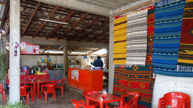 Restaurant, Teotitlán del Valle. This canteen-style restaurant is typical of peri-urban areas such as Teotitlán del Valle, a village located some 20 kilometers from the center of Oaxaca City. The restaurant is simply but elegantly designed, with an open-air plan that takes advantage of cross-breezes during the hot days, and a sloped roof for drainage during the rainy season. The kitchen in the back serves double-duty for both the restaurant and the household. The proprietor belongs to a rug weaving cooperative, and displays their creations on the walls. An open air restaurant typical of the peri-urban landscape of Oaxaca with cement floors, red, plastic tables, and rugs hanging from the walls