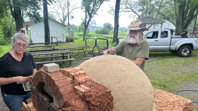 ARK oven collaborator Whitey Flagg and filmmaker Skye Borgman work on the sand form for the oven dome. Clay mined from the site, along with Kernza straw baled from the previous year Kernza harvest, will be combined and then packed around insulating beer bottles to form the walls of the oven. Photo by the author, 2022. Two people standing around a clay oven