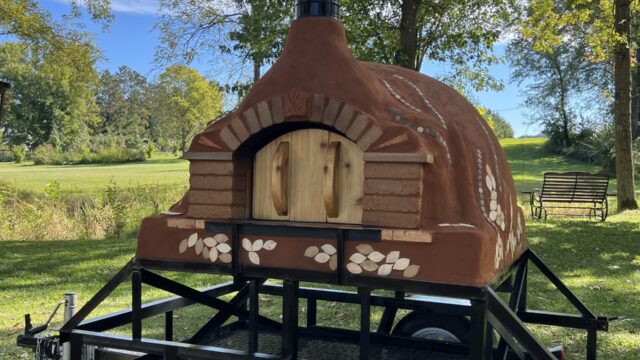 ARK oven, built from the clay of the ARK site and fired up at the 2022 DTour to bake Kernza crackers and shortbread from this year's ARK Kernza harvest. Photo by the author, 2022. Clay and brick oven with decorative prints and wood door