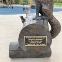 Bench Dedication This side view of the bronze bench bears a plaque that reads: "Gifted to the residents of Mountain Shadows by creators and sponsors of the 'Faces of the Fire' exhibit". The playground is visible in the background.