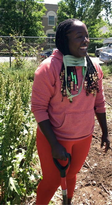 Woman wearing red pants and pink sweatshirt holds a shovel and smiles next to a plant.